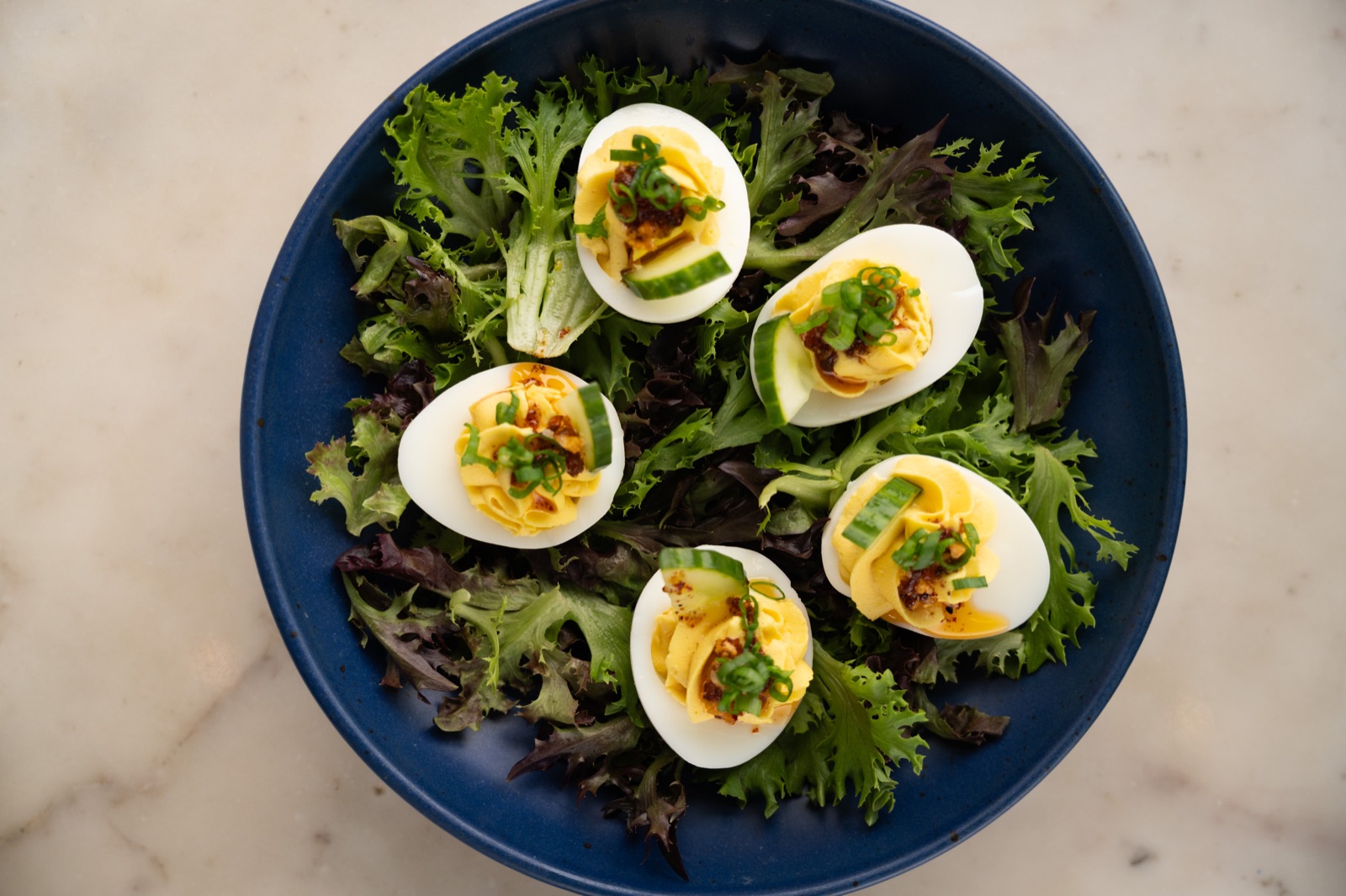Deviled eggs arranged over greens in a dark blue bowl, photographed from above.